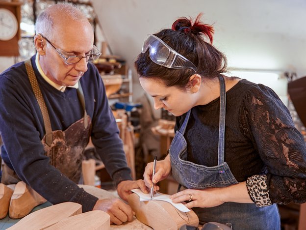 A woman traces a line on top of a piece of wood in the shape of a shoe. An older man helps hold the piece of wood. They both wear aprons and safety glasses.
