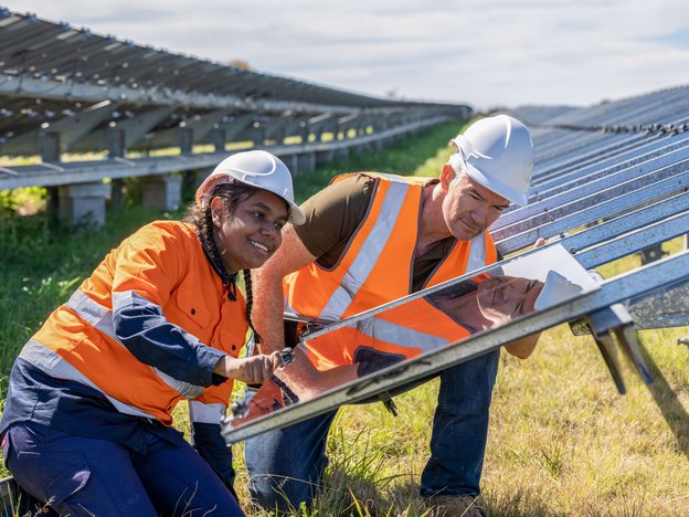 A woman and a man in orange and navy uniforms and white helmets kneel in front of solar panels in a solar farm.