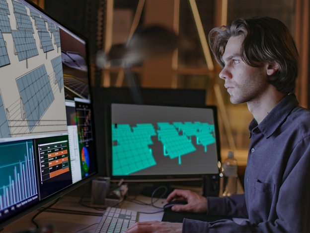 A man sits at a desk using a mouse and keyboard in front of two computer monitors. The monitors display solar panels.