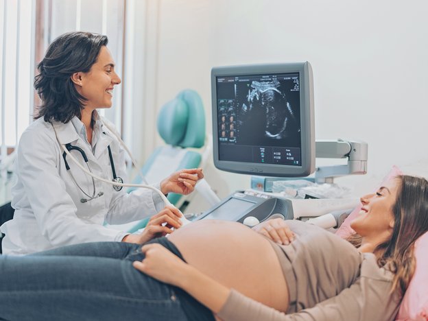 A female sonographer in a white lab coat does an ultrasound scan on a pregnant patient. The patient is lying on a hospital bed as the sonographer guides a scanning tool over her stomach and observes a computer screen displaying the sonogram.