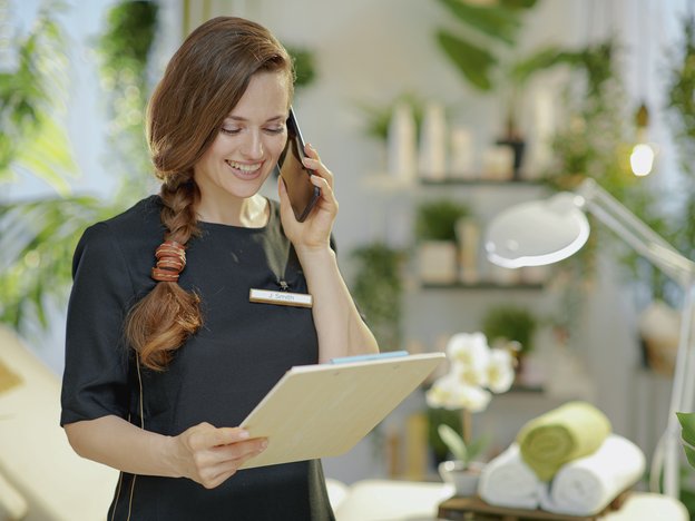 A woman holds a mobile phone to her ear in one hand and a notepad in the other. She wears a black uniform shirt.