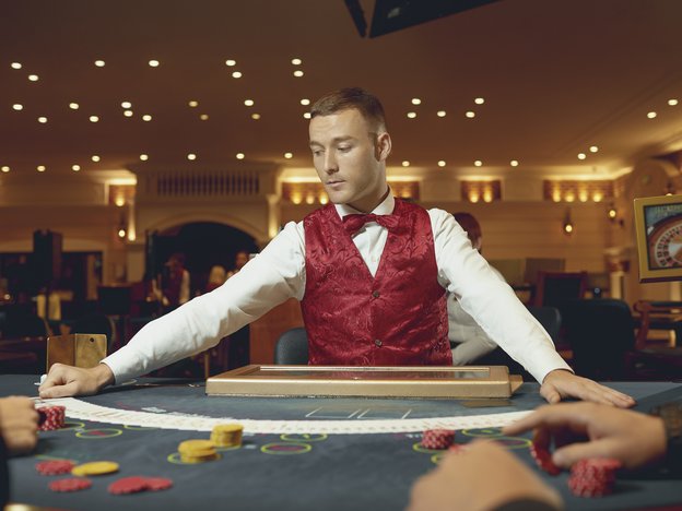 A man looks at a line of cards on a poker table. He wears a red vest, red bowtie and a white shirt.