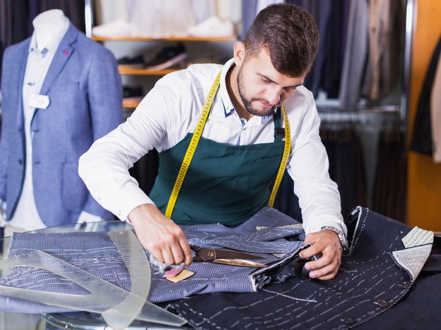 A man cuts through clothing fabric. He wears an apron and there is measuring tape around his neck.