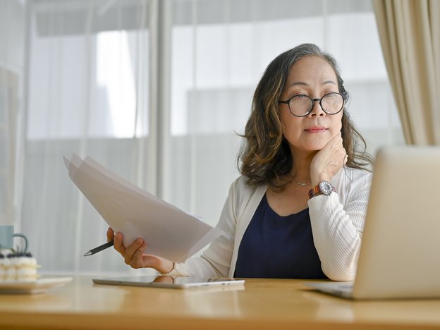 A woman wearing glasses sits at a desk. She holds a pen and paper and looks at a laptop.