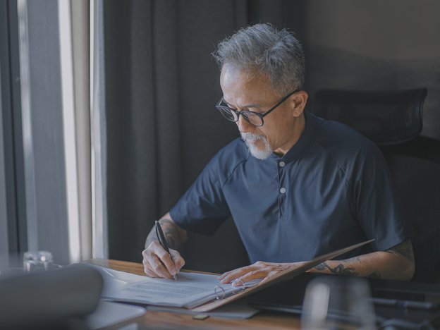 A man in a navy polo shirt sits at a desk writing on a notepad in a folder.
