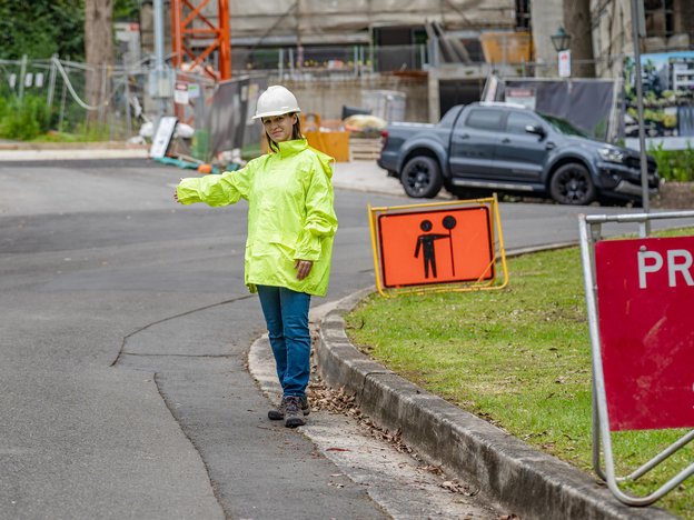 A woman gestures with her right hand to the right direction. She wears a green high visibility jacket and a white helmet.