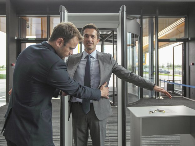 A man frisks another man at a security check in. There is a millimeter wave scanner behind them. They both wear suits.