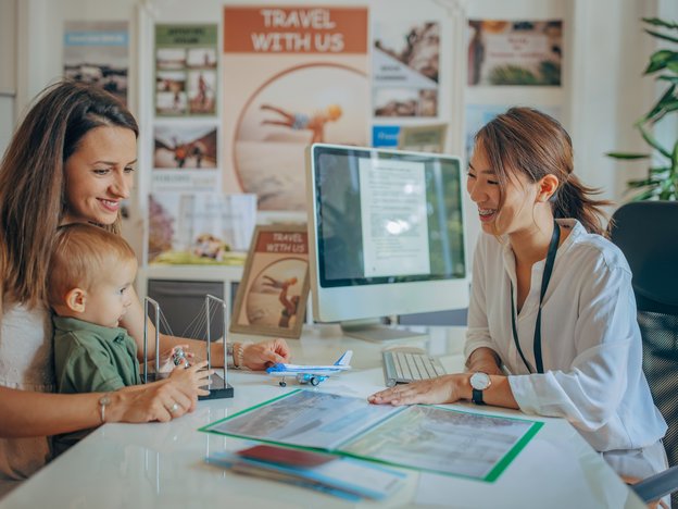 A female travel agent shows a mother and her baby some documents in folder. They are sitting opposite each other at a desk in an office. Behind them are tourism posters and a computer monitor.