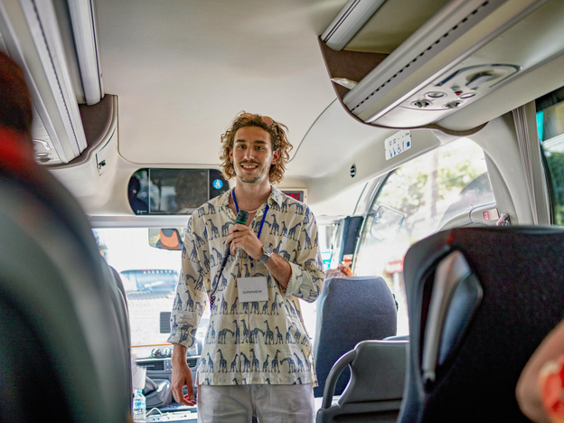 A man holds a microphone and stands inside a vehicle. He wears a lanyard with a tag that says 'Supervisor'.