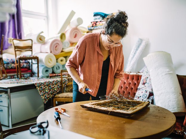 A person adds springs to a chair seat. They are surrounded by furniture, fabric and other material.