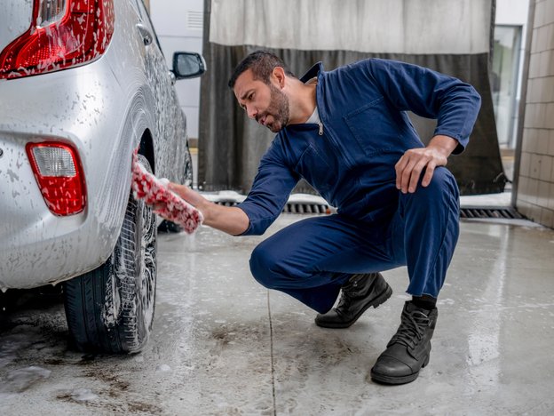 A man cleans a silver car. They are wearing blue overalls and black boots.