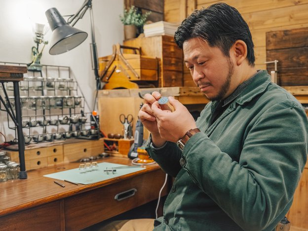 A man in a green shirt with short black hair sits at a desk making a watch with tools surrounding him in a wooden room.