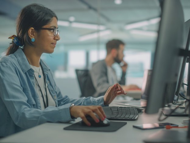 A woman uses a mouse and keyboard and looks at her computer at a desk. She is in an office sitting next to a male colleague.