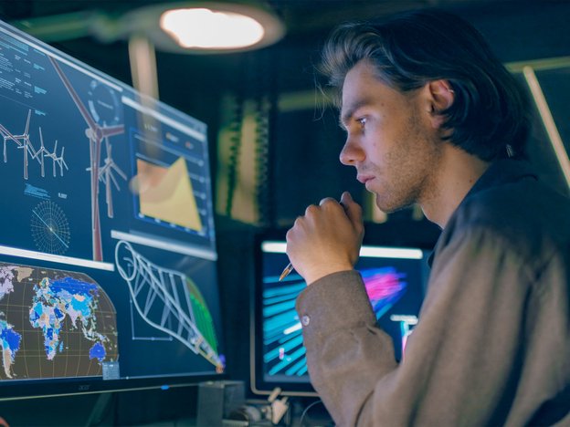 A man observes two computer monitors displaying wind movements. He is holding a pen.