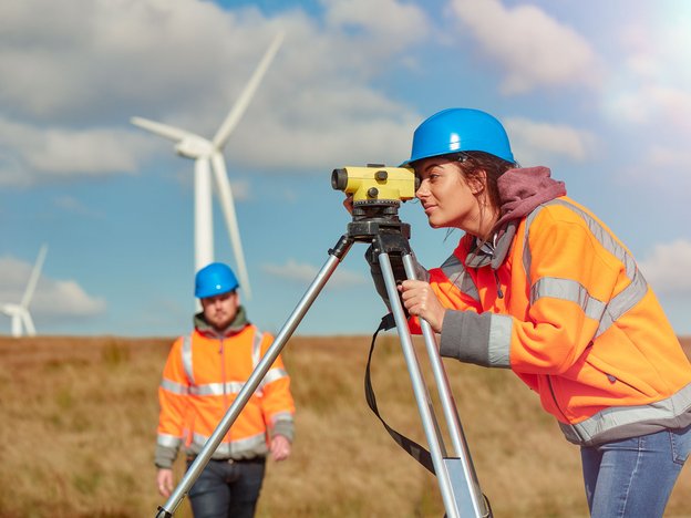A woman looks through a yellow vision device. A man stands behind her in a wind farm field. They are both wearing orange jackets and blue helmets.