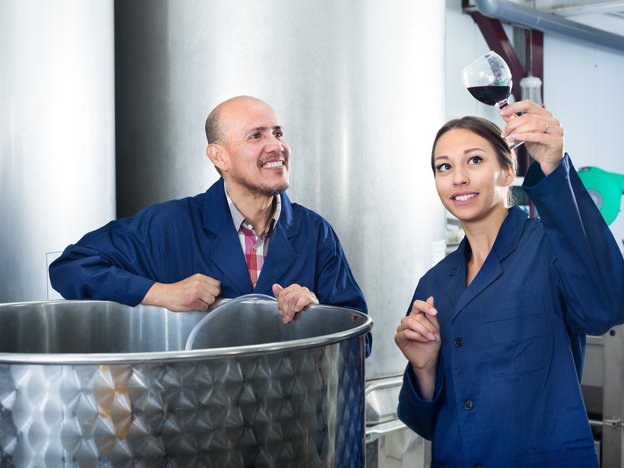 Two winery workers observe a glass of wine. They are in a standing next to steel barrels.