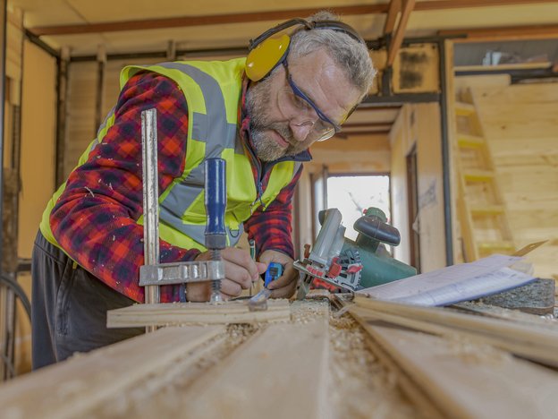 A man cuts some wood on a table using a cutting machine. He wears a yellow fluorescent vest, safety glasses and yellow earmuffs.