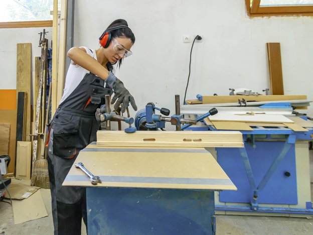 A woman cuts some wood on a table using a cutting machine in a workshop. She wears safety glasses, red earmuffs and a grey apron.