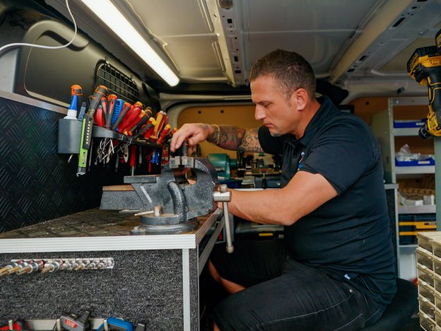 A locksmith works at their workbench. They are sitting in the back of a van, surrounded by tools and equipment.