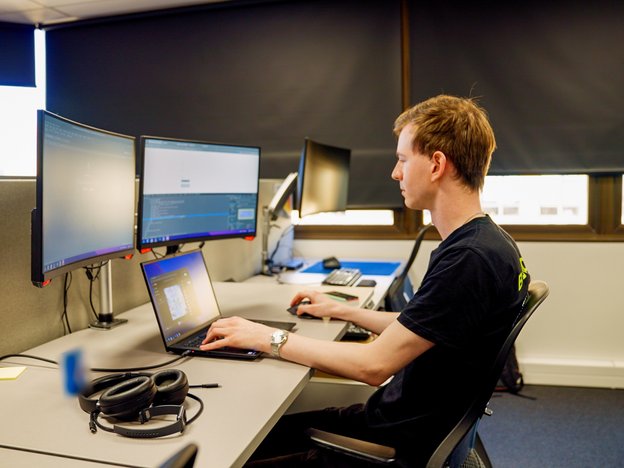 A person sits at their desk on their laptop, which is connected to two monitors. They are in an office.
