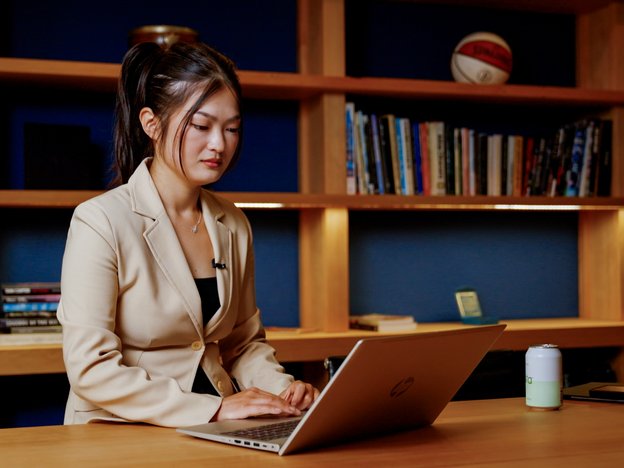 A person sits at an office desk using a laptop. Behind them is a bookshelf.