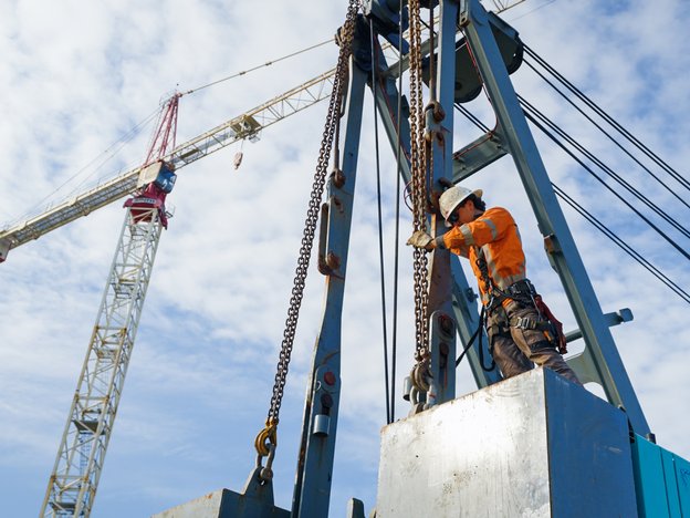 A worker operates a rig. They are wearing a safety helmet, an orange high-vis top and work pants with a safety harness.