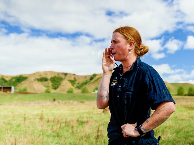 A farmer blows a whistle. They are wearing overalls, standing in a field