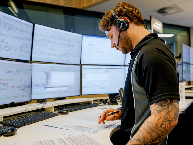 A person looks at plans on desk. They are standing in a control room, wearing a headset.