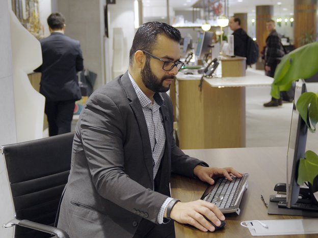 A man sits on a chair and types on a keyboard. He looks at a computer screen.
