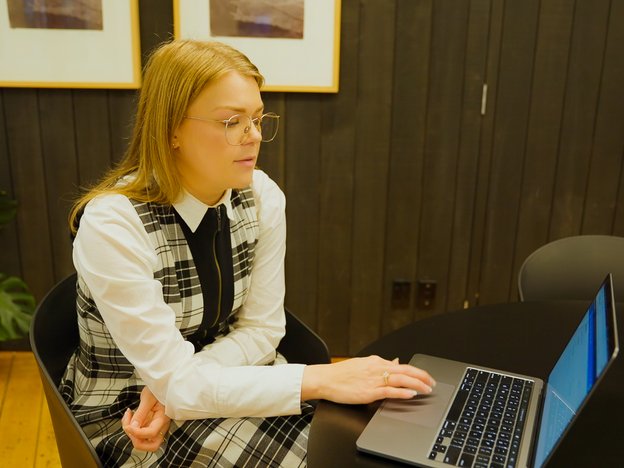 A woman wearing a white and black checkered dress with blonde hair sits at a desk with a laptop in an office.