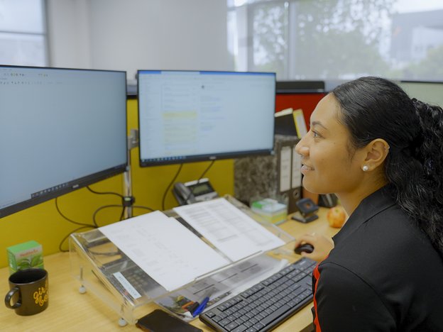 A young smiling woman wearing black sits at a desk in front of two monitors, a keyboard, documents and other office stationery. She is in an office.
