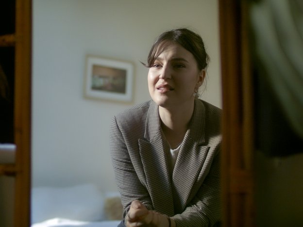 A person looks into a mirror. They are sitting on a bed wearing a brown coat and have a concerned expression.