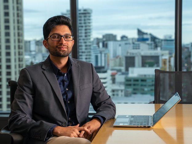 A young man sits in a modern office in a suit. His laptop is open on the desk.