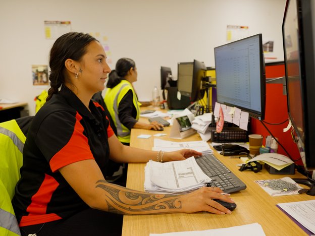 Woman with black hair sits in front of a computer monitor using a mouse and keyboard. She wears a black and red shirt, and a fluorescent yellow vest drapes over the back of her desk chair. A woman sits behind her in a yellow vest.