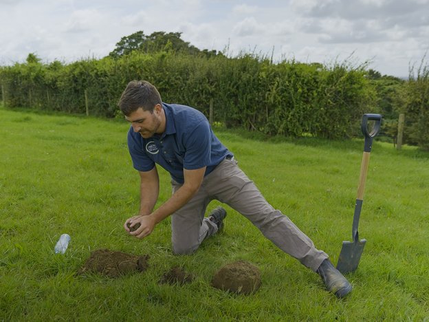 A man leans on one knee with his other foot outstretched on a grass paddock. He has dug a hole with a shovel and is molding dirt with his hands.