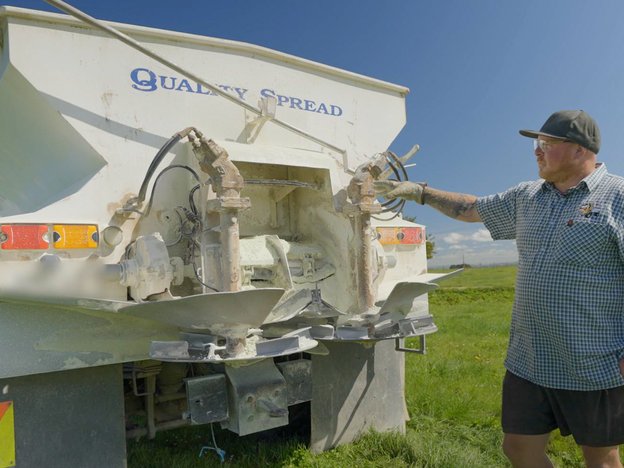 A man in a blue and white checkered shirt, brown shirts and gloves stands behind a large truck in a field of grass.