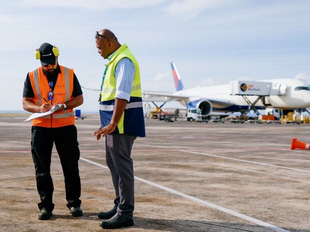 Two men in high-visibility vests stand on an airport tarmac with a plane in the background.