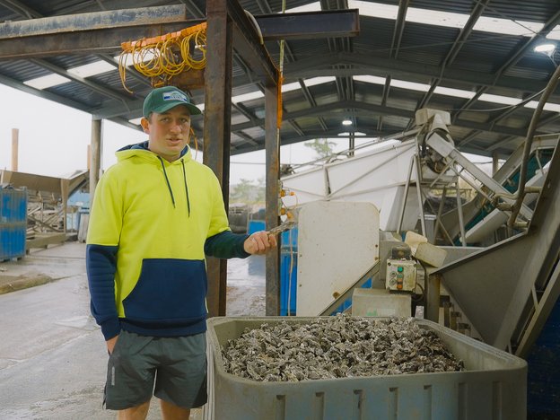A man wearing a fluro yellow and dark blue sweatshirt, a green cap and dark green shorts stands next to a large container of oysters outdoors.