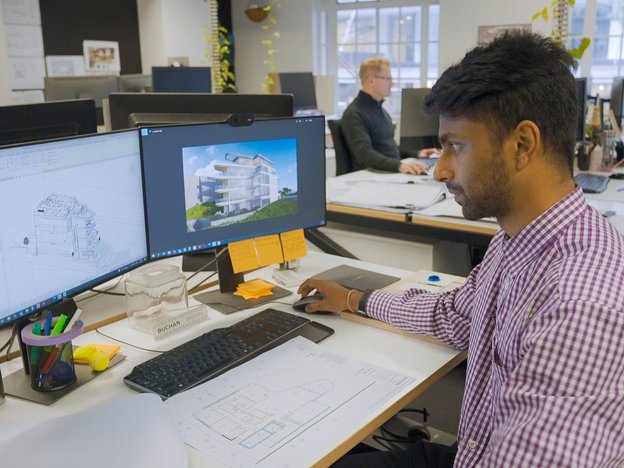 A young man designs a building on his computer, he sits in an office space. A colleague sits at another desk.