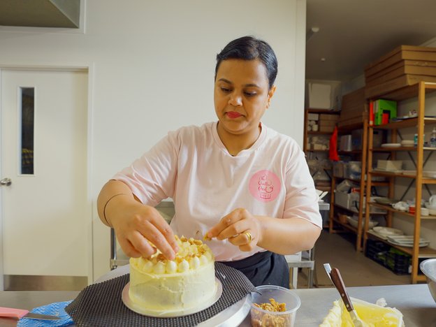 A woman in a pink shirt decorates and ices a white cake in a kitchen. She is surrounded by baking ingredients and shelves with banking equipment.