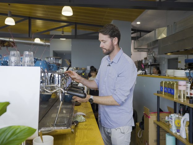 Man in blue shirt and beige trousers grinds coffee at a machine in a cafe.