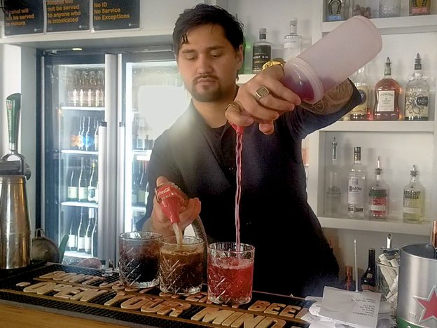 A bartender pours ingredients into glasses. They are behind the bar pouring soda into one glass, while pouring liquid flavouring into the other.