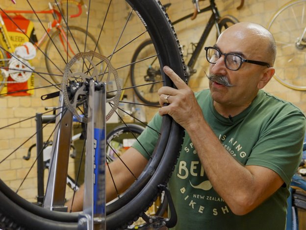 Man with a moustache wears glasses and a green t-shirt. He works on a bicycle wheel and spokes in a bicycle factory.
