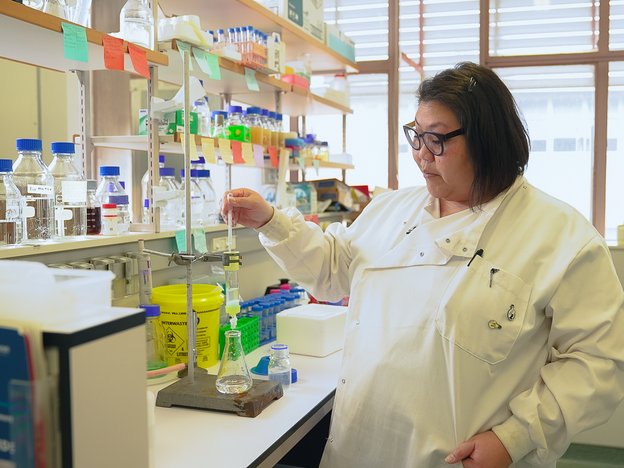 A woman in a lab coat drips solution into a beaker using a pipette. Behind her are many test tubes and labelled bottles.