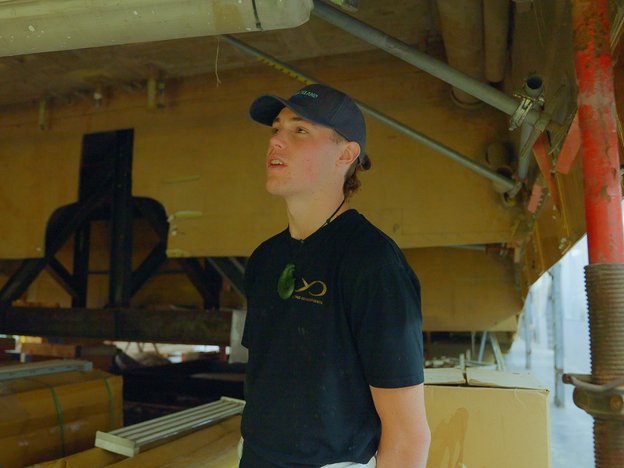 A young builder stands underneath the hull of a boat in construction. They are surrounded by timber and boxes.