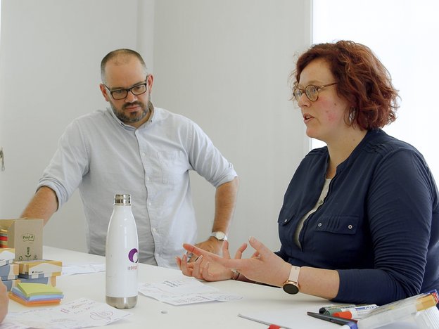Two people gather around a table in discussion. The man stands while the woman sits and speaks. There are pieces of paper, post-it notes and a drink bottle in front of them.