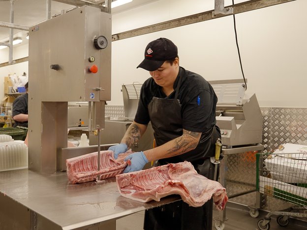 A man uses a machine to cut meat. He is wearing a black apron and blue gloves.