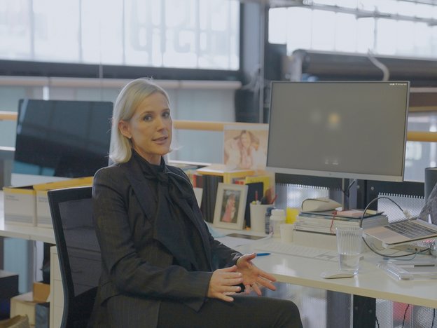 A businesswoman in a suit sits in an open office desk. Their desk has a computer, a laptop and other office supplies.