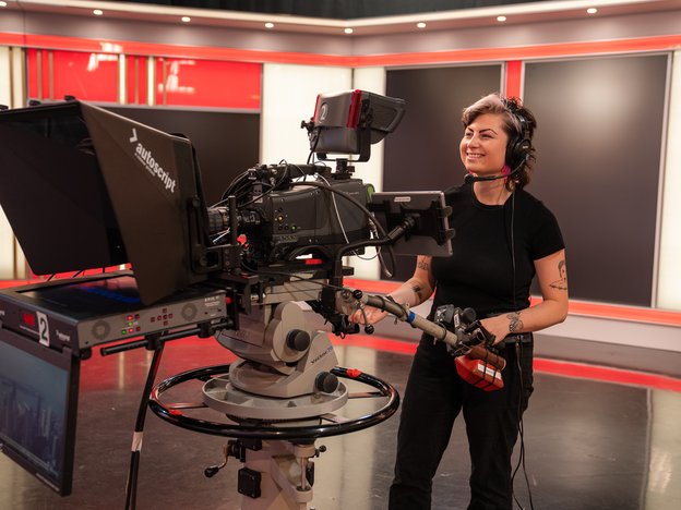 A camera operator holds a video camera and teleprompter stand. They are dressed in all black, in a news studio.