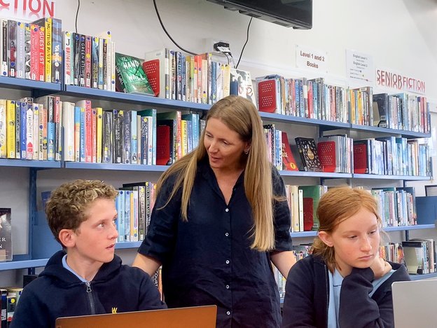 A woman stands in a school library helping two young students.